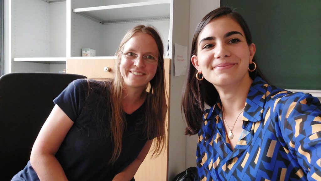 A selfie of Lisa Sauermann and Raffaella Mulas. They are sitting on a desk with a blackboard in the background, and they are looking into the camera, smiling.